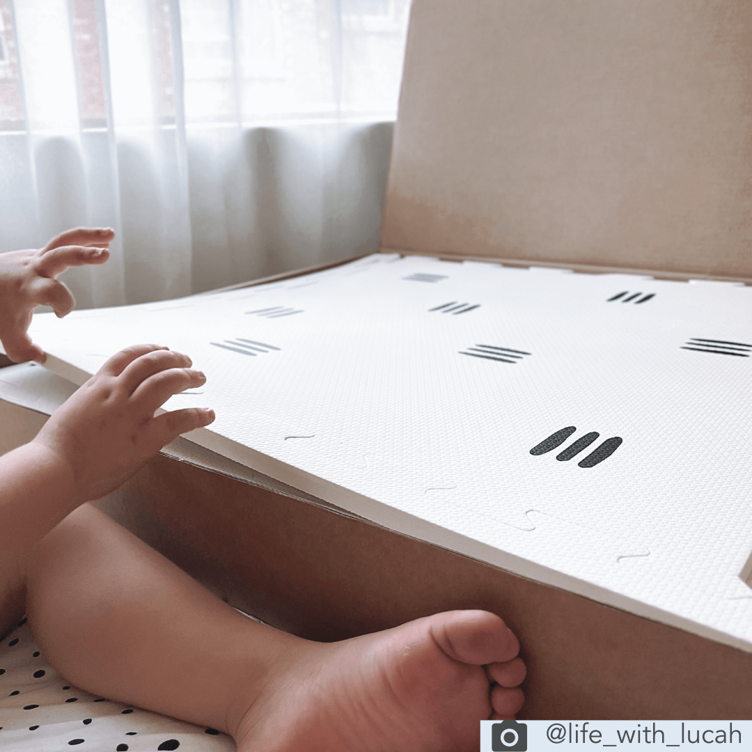 This image showcases the White with black stripes play mat, as it is unboxed by a child. This play mat features a sleek and modern monochrome design of white mat with 3 black stripes. The unique and timeless design of this play mat adds a touch of class and sophistication to any play area or nursery.